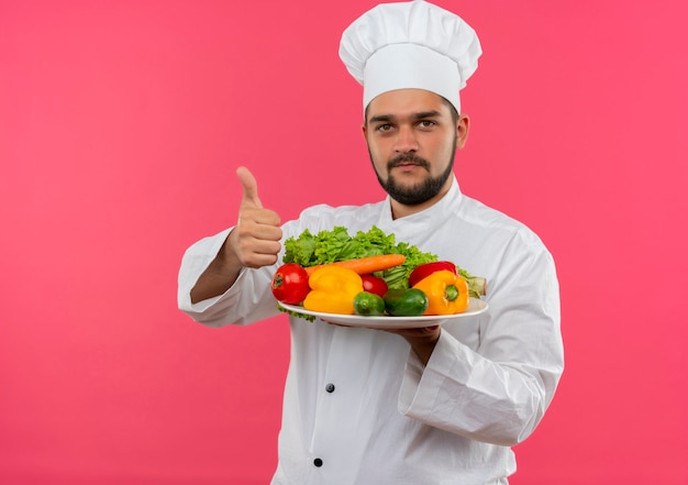 野菜のプレートを保持し、ピンクのスペースで隔離の親指を見せてシェフの制服を着た若い男性料理人を喜ばせる