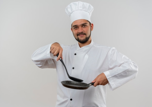 Pleased young male cook in chef uniform holding slotted spoon and frying pan isolated on white space