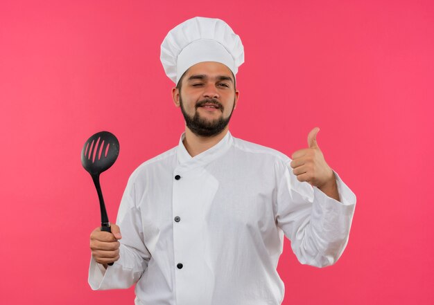 Pleased young male cook in chef uniform holding spatula and showing thumb up and winking isolated on pink space