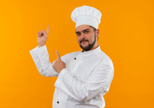Pleased young male cook in chef uniform pointing up isolated on orange space 