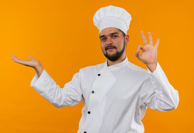空の手を示し、オレンジ色のスペースで隔離のokサインをしているシェフの制服を着た若い男性料理人を喜ばせる