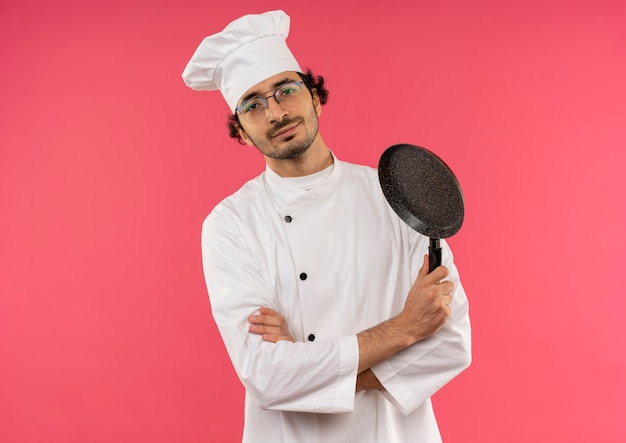 Pleased young male cook wearing chef uniform and glasses crossing hands and holding frying pan on pink