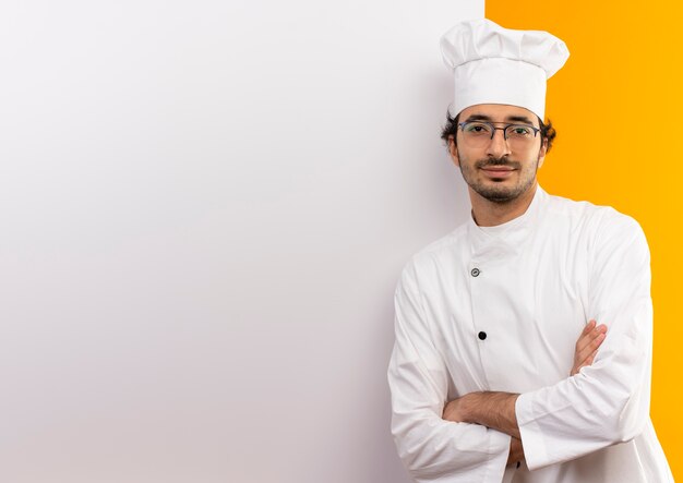 Pleased young male cook wearing chef uniform and glasses crossing hands standing white wall isolated on yellow wall with copy space