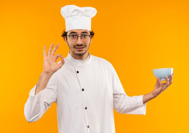 Pleased young male cook wearing chef uniform and glasses holding bowl and showing okey gesture isolated on yellow wall