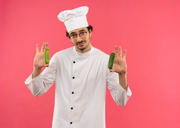Довольный молодой мужчина-повар в униформе шеф-повара и очках держит огурец на розовой стене