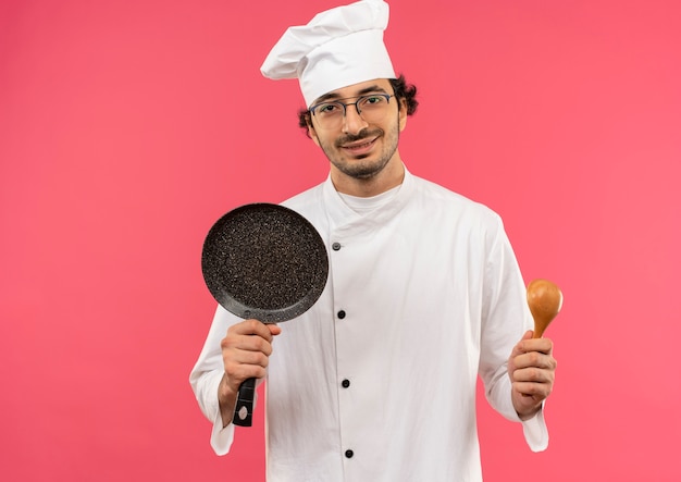 Довольный молодой мужчина-повар в униформе шеф-повара и в очках держит сковороду и ложку