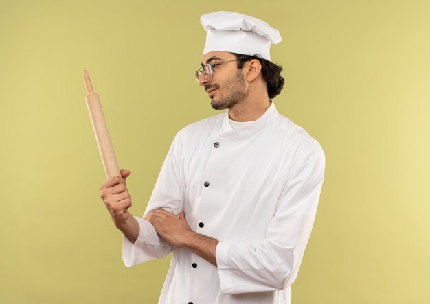 Pleased young male cook wearing chef uniform and glasses holding and looking at rolling pin isolated on green wall