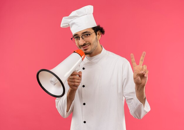 Довольный молодой мужчина-повар в униформе шеф-повара и очках держит громкоговоритель вокруг рта и демонстрирует мир, изолированный на розовой стене