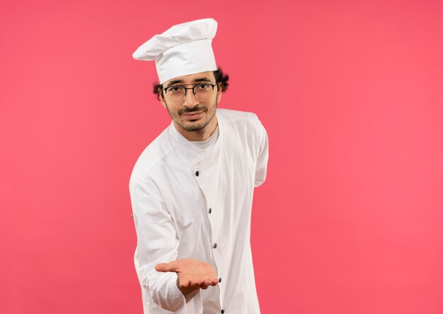 Pleased young male cook wearing chef uniform and glasses holding out hand isolated on pink wall