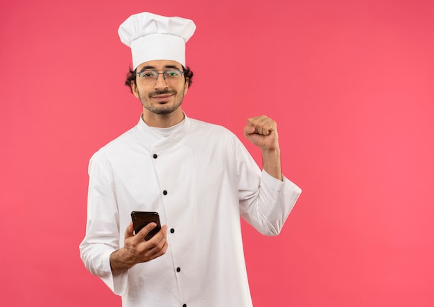 Pleased young male cook wearing chef uniform and glasses holding phone and showing yes gesture isolated on pink wall