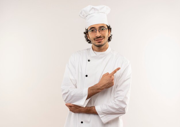 Pleased young male cook wearing chef uniform and glasses points to side isolated on white wall with copy space