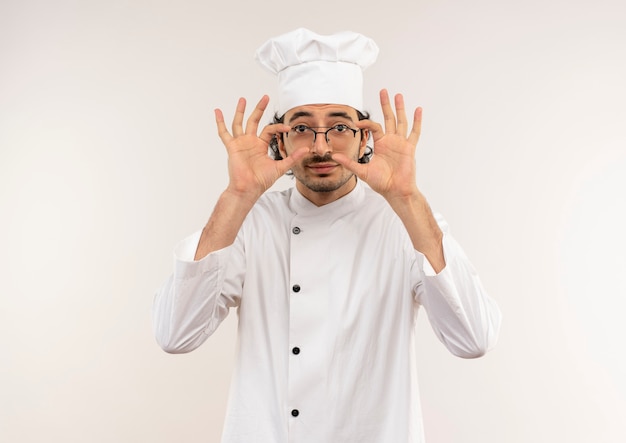 Pleased young male cook wearing chef uniform and glasses putting hand on glasses isolated on white wall