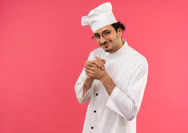 Pleased young male cook wearing chef uniform and glasses showing handshakes isolated on pink wall