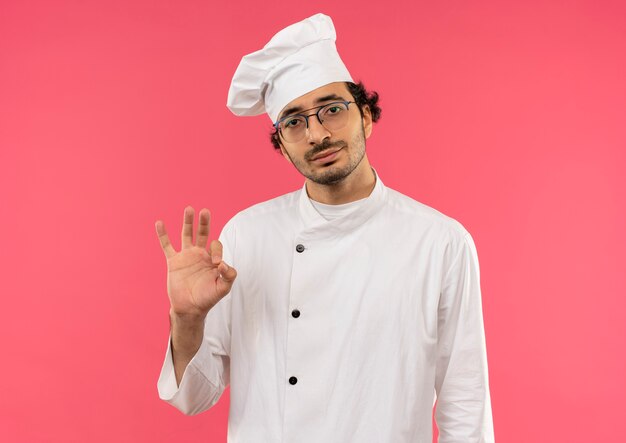 Pleased young male cook wearing chef uniform and glasses showing okey gesture isolated on pink wall