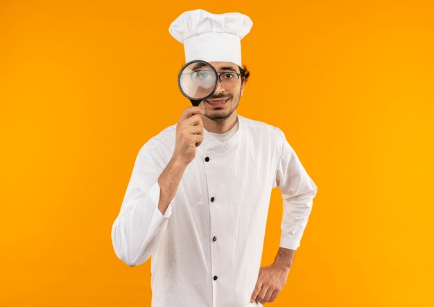 Pleased young male cook wearing chef uniform and glasses with magnifier isolated on yellow wall