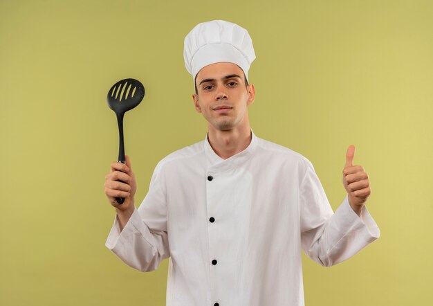 Pleased young male cook wearing chef uniform holding spatula his thumb up 