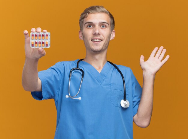 Pleased young male doctor wearing doctor uniform with stethoscope holding pills spreading hand isolated on orange wall