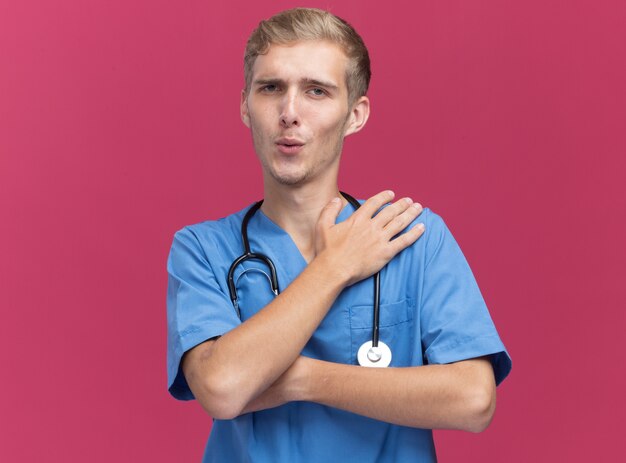 Довольный молодой мужчина-врач в униформе врача со стетоскопом, положив руку на плечо, изолированную на розовой стене