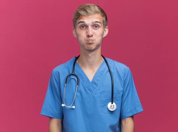 Pleased young male doctor wearing doctor uniform with stethoscope showing kiss gesture isolated on pink wall