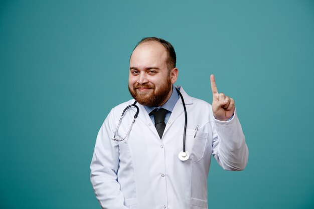 Pleased young male doctor wearing medical coat and stethoscope around his neck looking at camera pointing up isolated on blue background