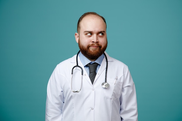 Felice giovane medico maschio che indossa cappotto medico e stetoscopio intorno al collo guardando il lato isolato su sfondo blu