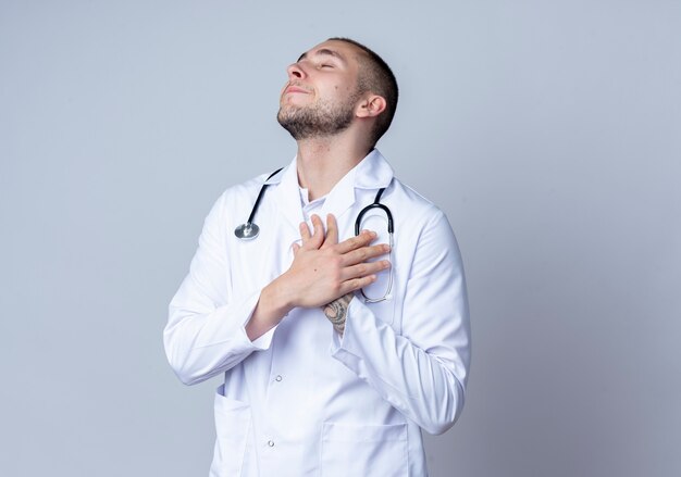 Pleased young male doctor wearing medical robe and stethoscope around his neck putting hands on his heart with closed eyes isolated on white wall