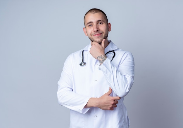 Pleased young male doctor wearing medical robe and stethoscope around his neck touching his chin and elbow isolated on white wall