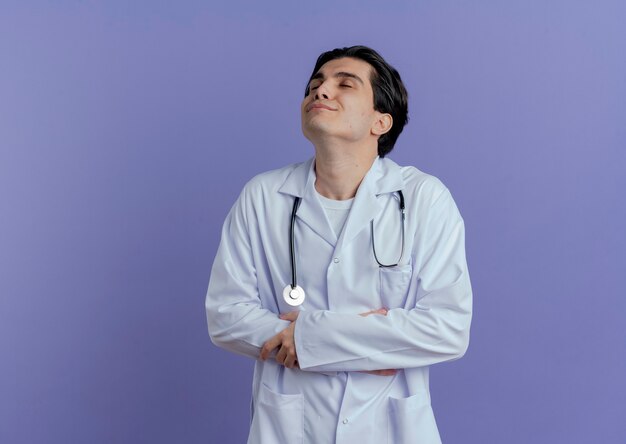 医療用ローブと聴診器を身に着けている若い男性医師は、コピースペースのある紫色の壁に隔離された目を閉じて腹と腕に手を置いて喜んでいます