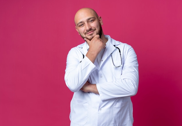 Pleased young male doctor wearing medical robe and stethoscope putting hand on chin