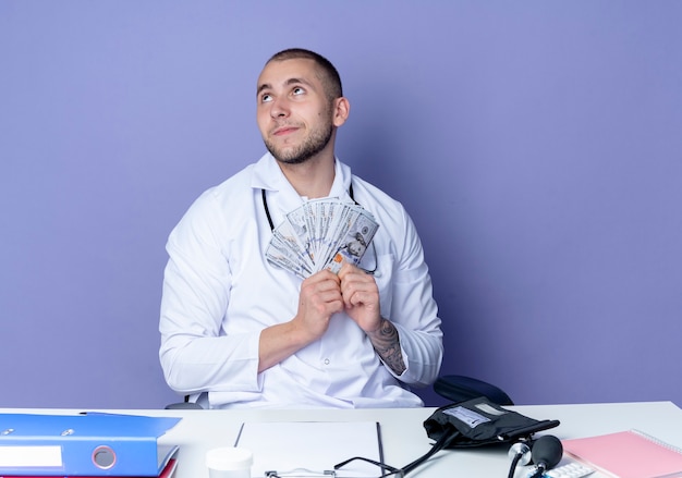 Free photo pleased young male doctor wearing medical robe and stethoscope sitting at desk with work tools holding money and looking up isolated on purple wall