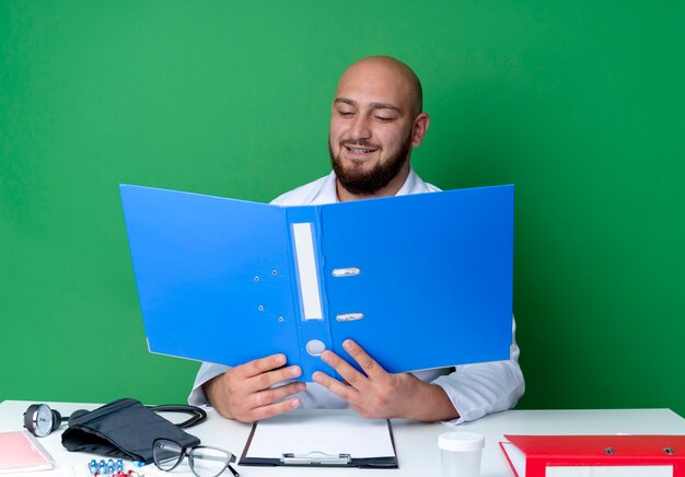 Pleased young male doctor wearing medical robe and stethoscope sitting at desk