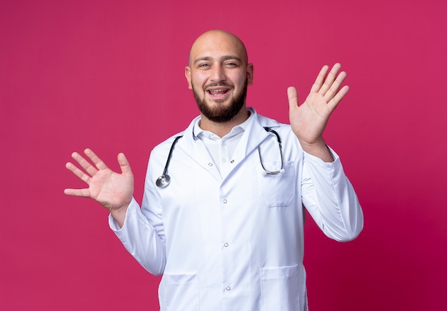 Pleased young male doctor wearing medical robe and stethoscope spreads hands isolated on pink