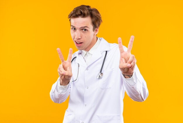 Pleased young male doctor wearing medical robe with stethoscope showing peace gesture isolated on orange wall