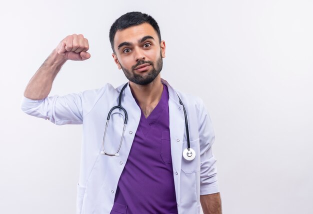 Pleased young male doctor wearing stethoscope medical gown doing strong gesture on isolated white background