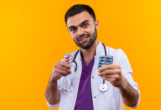 Free photo pleased young male doctor wearing stethoscope medical gown holding pills on isolated yellow background