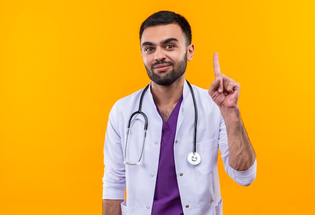 Pleased young male doctor wearing stethoscope medical gown points to up on isolated yellow background