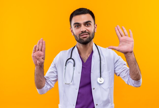 Pleased young male doctor wearing stethoscope medical gown showing different numbers on isolated yellow background
