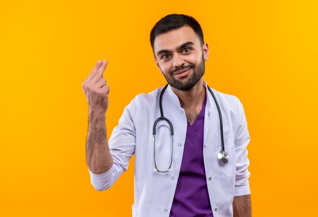 pleased young male doctor wearing stethoscope medical gown showing tip gesture on isolated yellow wall