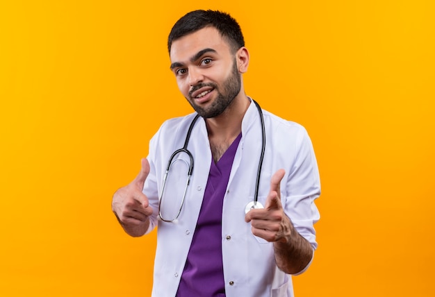 Довольный молодой мужчина-врач в медицинском халате со стетоскопом показывает вам жест на изолированном желтом фоне