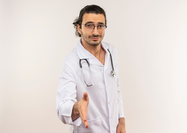 Pleased young male doctor with optical glasses wearing white robe with stethoscope holding out hand to on isolated white wall with copy space