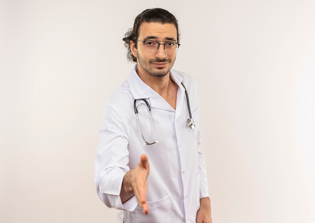 Pleased young male doctor with optical glasses wearing white robe with stethoscope holding out hand to on isolated white wall with copy space