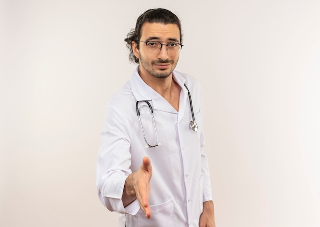 Довольный молодой мужчина-врач в оптических очках, одетый в белый халат со стетоскопом, протягивает руку к изолированной белой стене с копией пространства