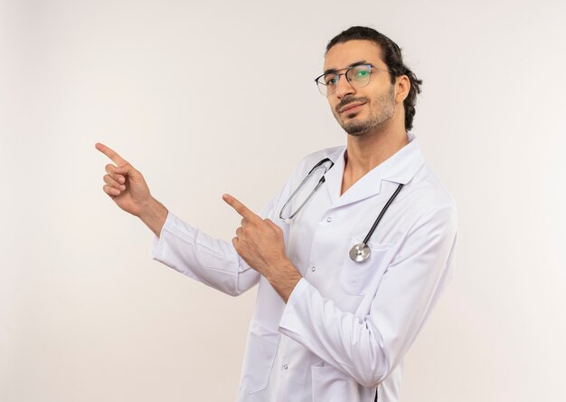 Pleased young male doctor with optical glasses wearing white robe with stethoscope points to side on isolated white wall with copy space