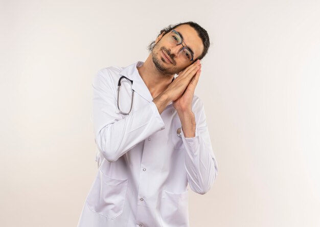 Pleased young male doctor with optical glasses wearing white robe with stethoscope showing sleep gesture on isolated white wall with copy space