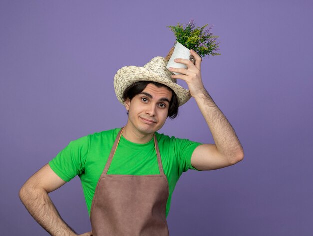 Pleased young male gardener in uniform wearing gardening hat holding flower in flowerpot on head