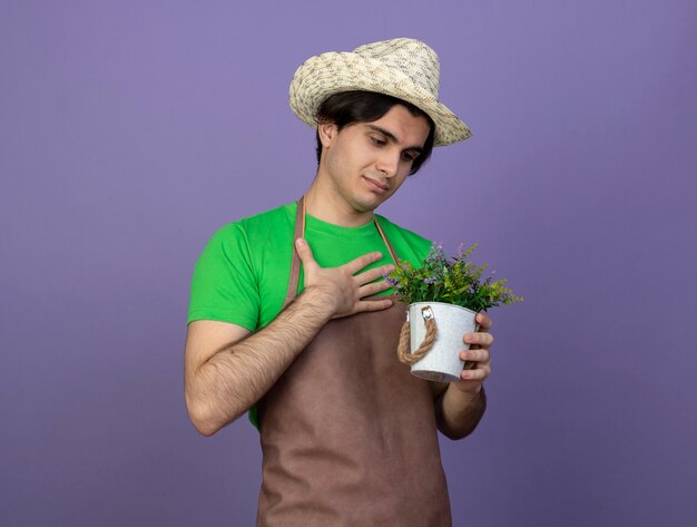 Pleased young male gardener in uniform wearing gardening hat holding and looking at flower in flowerpot putting hand on chest