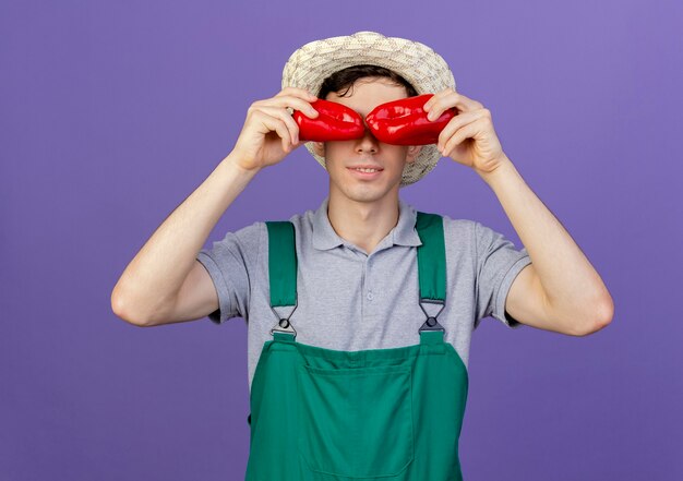Pleased young male gardener wearing gardening hat covers eyes with peppers 