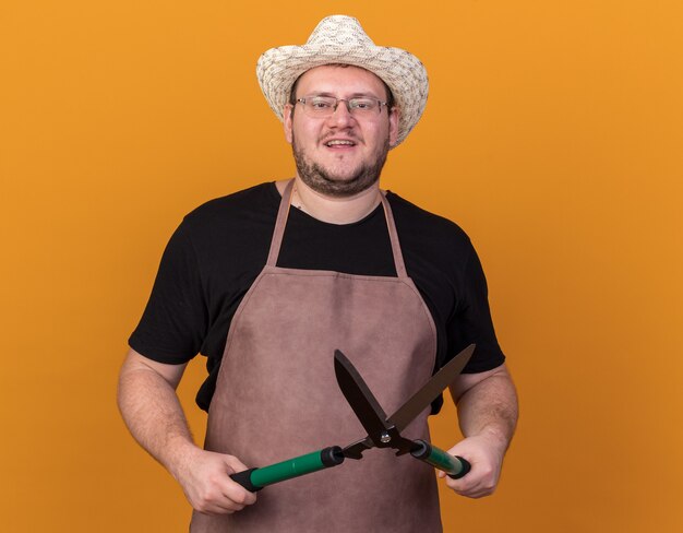 Pleased young male gardener wearing gardening hat and gloves holding clippers isolated on orange wall