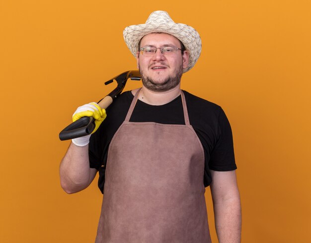Довольный молодой мужчина-садовник в садовой шляпе и перчатках держит лопату на плече, изолированную на оранжевой стене