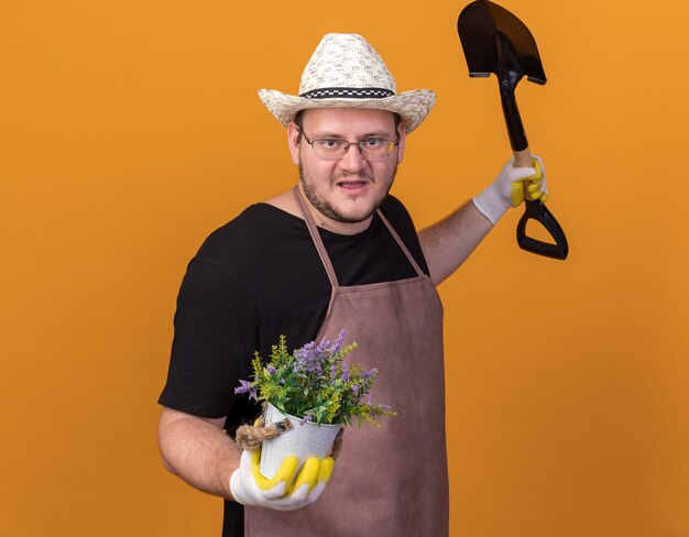 Довольный молодой мужчина-садовник в садовой шляпе и перчатках держит лопату с цветком в цветочном горшке, разводя руками, изолированными на оранжевой стене
